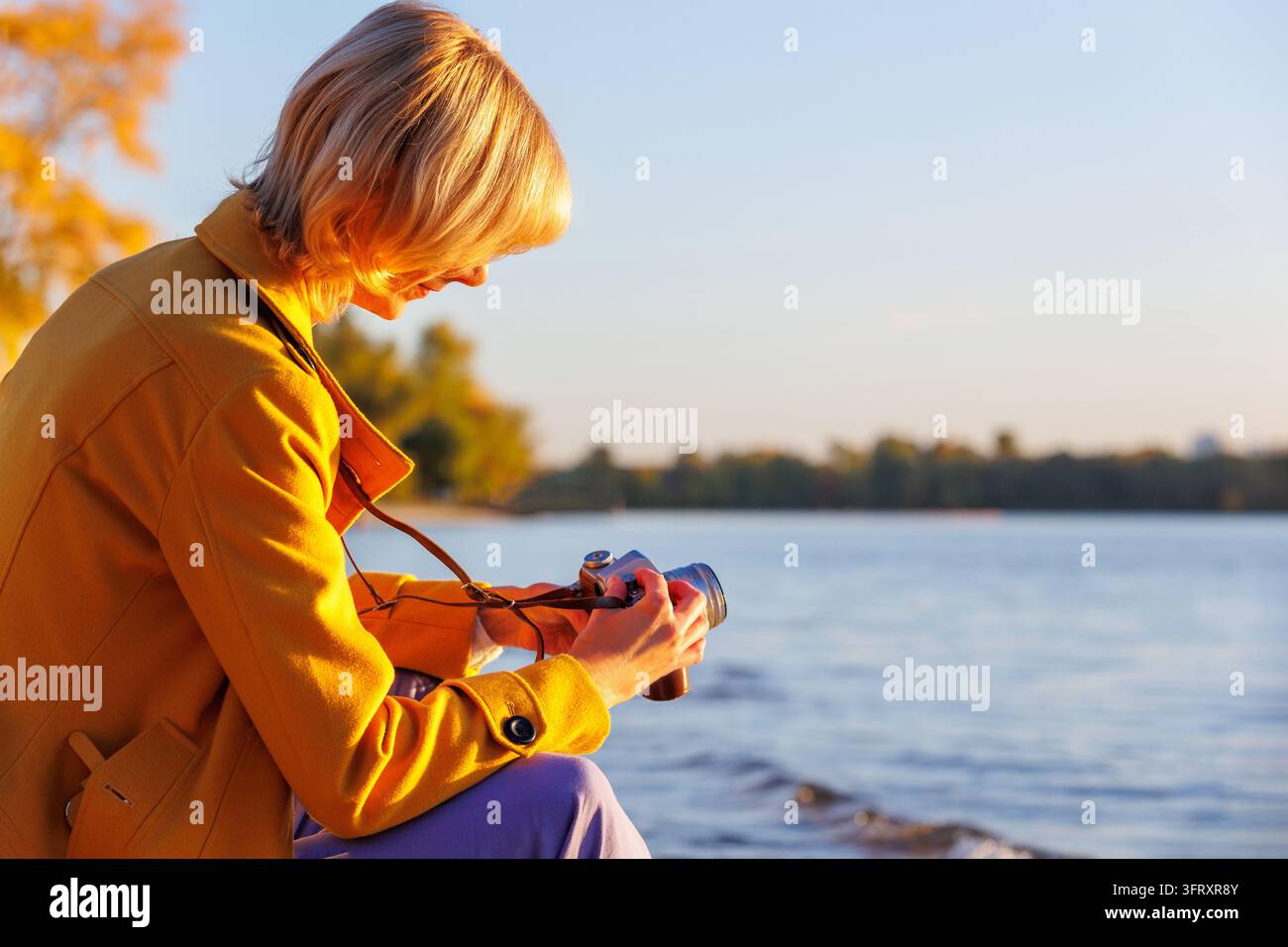 Donna seduta vicino al lago, regola i comandi di una fotocamera analogica vintage nella calda luce del tramonto. Hobby della fotografia analogica, tempo libero creativo, rilassamento consapevole, en Foto Stock