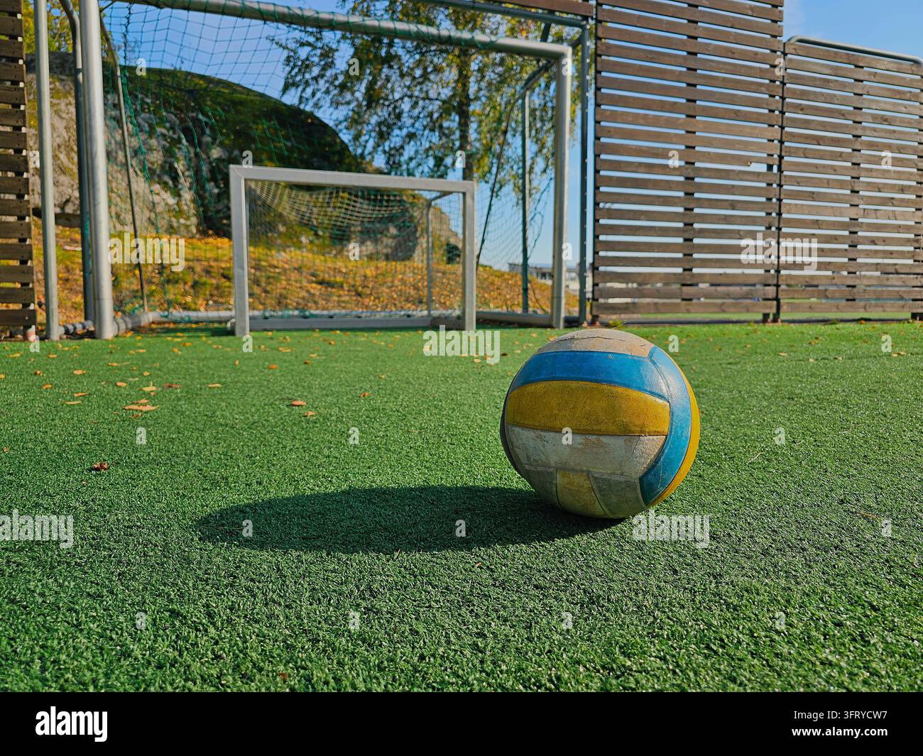 Una palla multicolor consumata poggia su erba artificiale verde davanti a un gol di calcio vuoto con rete. - Immagine stock catturata con smartphone