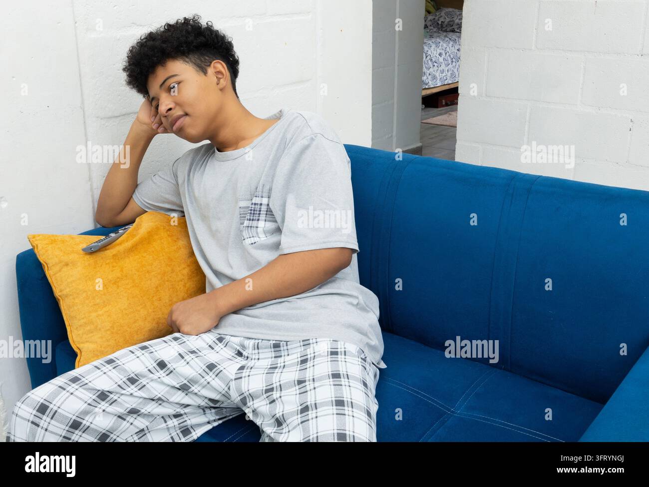 Adolescente dalla pelle scura seduto sul divano a guardare la televisione, con la mano appoggiata sulla testa e un'espressione divertita. Scena domestica quotidiana, d Foto Stock