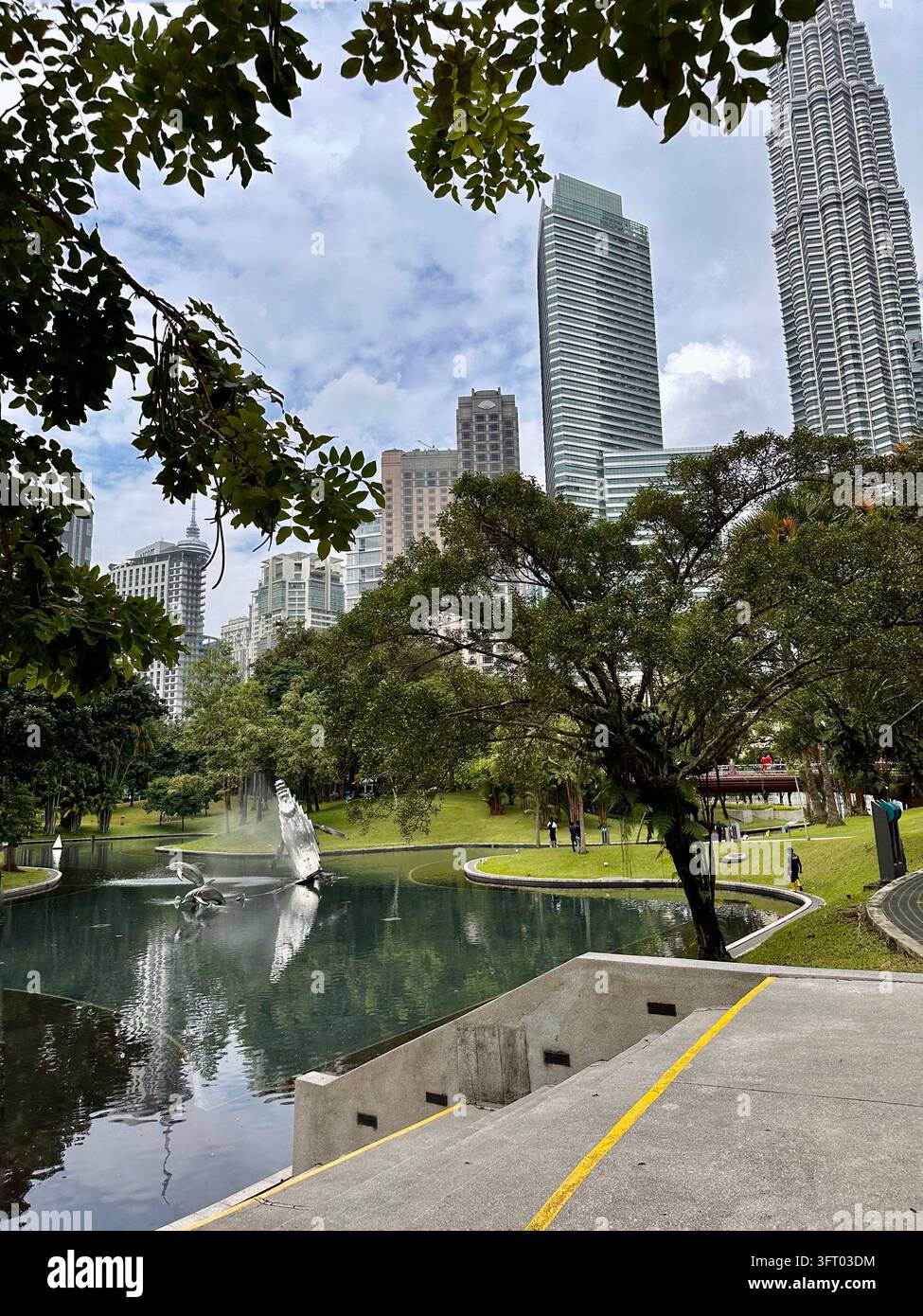 Kuala Lumpur, Malesia - 27 novembre 2022: Fontana scultorea si riflette tra alberi frondosi e grattacieli di Kuala Lumpur nella luce diffusa del giorno. - Immagine stock catturata con smartphone