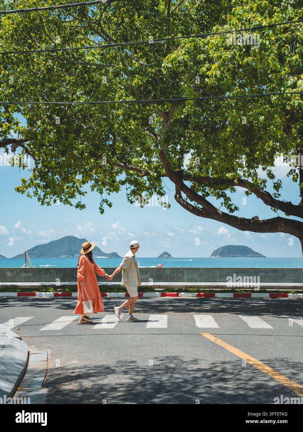 Coppia che si tiene per mano attraversa una strada costiera sotto una grande chioma di alberi con isole sullo sfondo, Con Dao, Vietnam Foto Stock