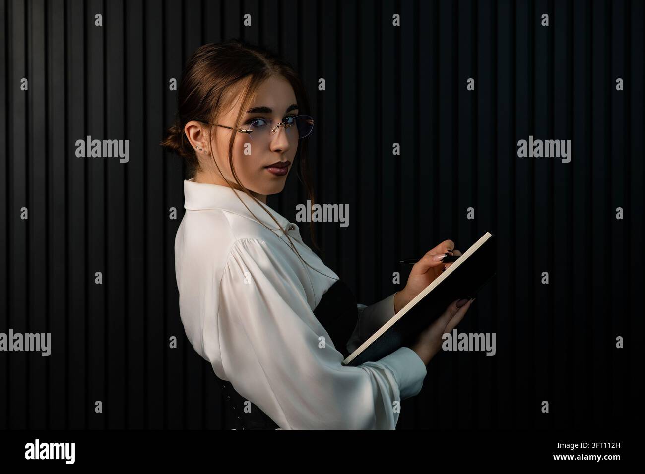 Giovane professionista sicuro di sé in camicia bianca che scrive su un taccuino, in piedi davanti a un muro moderno a strisce nere. Perfetto per il business e l'istruzione Foto Stock