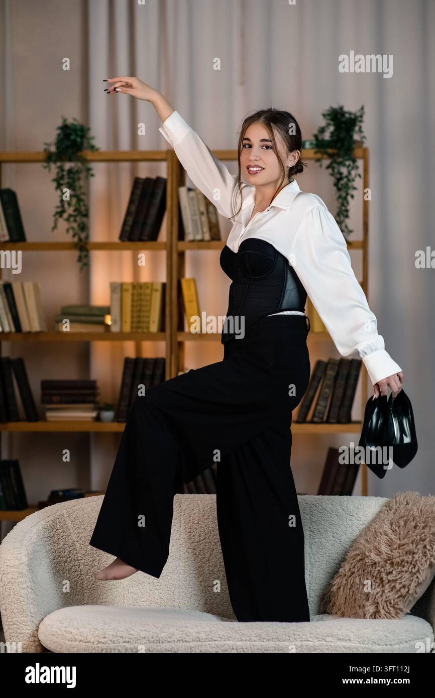 Giovane donna elegante in abito bianco e nero che posa su un divano moderno, tenendo delle scarpe, con scaffali di libri sullo sfondo. Foto Stock