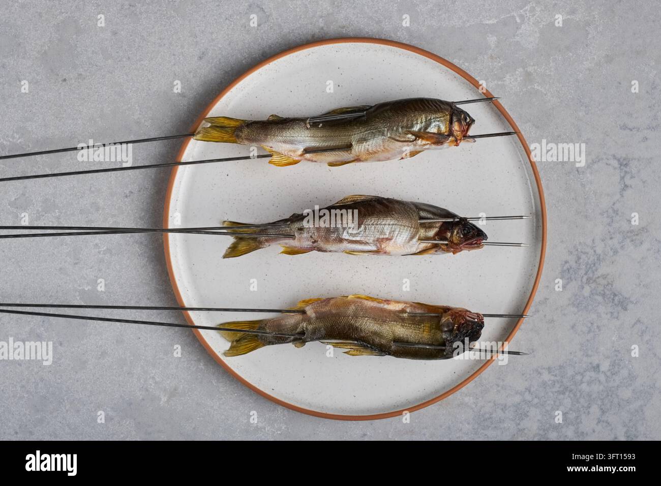 Tre piccoli pesci stagionati su spiedini di metallo su un piatto di ceramica, vista dall'alto Foto Stock