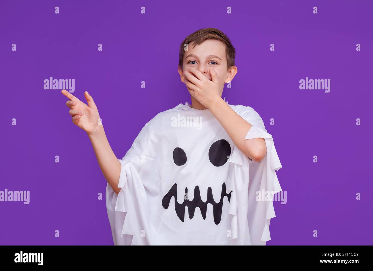 Ragazzo che indossa un costume da fantasma bianco con una faccia divertente che copre la bocca e punta un dito in modo giocoso. Foto Stock