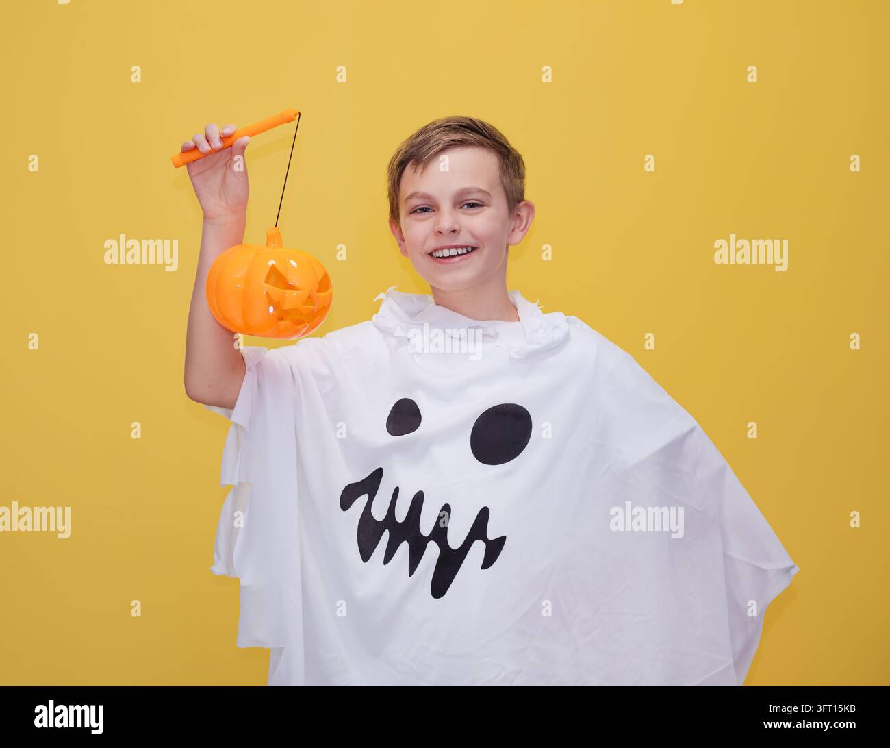 Ragazzo sorridente vestito da fantasma che tiene una lanterna di zucca arancione brillante, si diverte alla festa di Halloween con un'espressione allegra e un abbigliamento festoso. Foto Stock