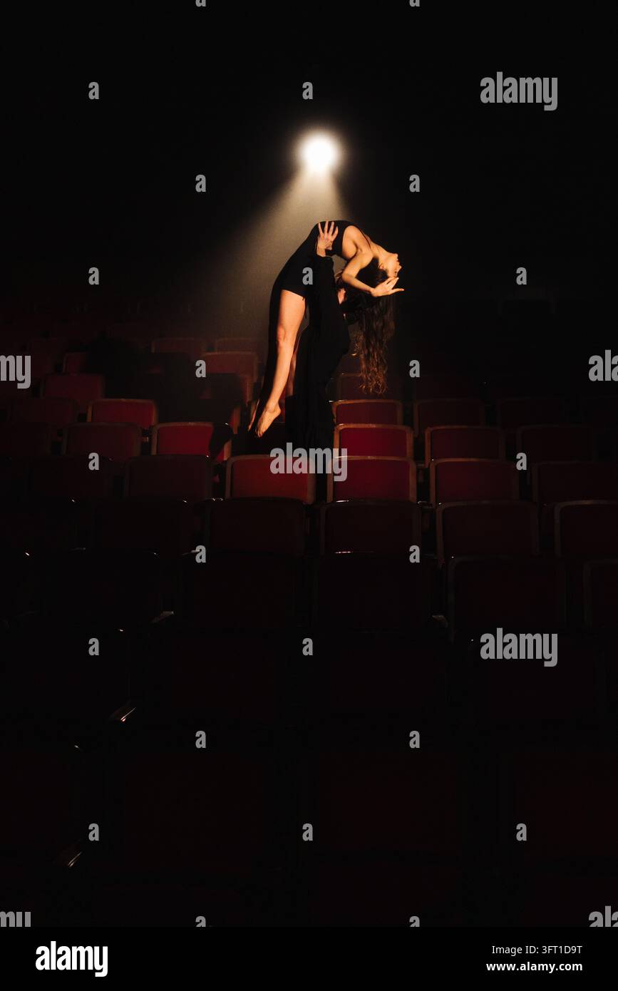 Sollevamento di danza contemporanea in un auditorium vuoto illuminato da un singolo riflettore che mette in mostra una ballerina arcuata sostenuta dal partner tra i sedili scuri del teatro Foto Stock