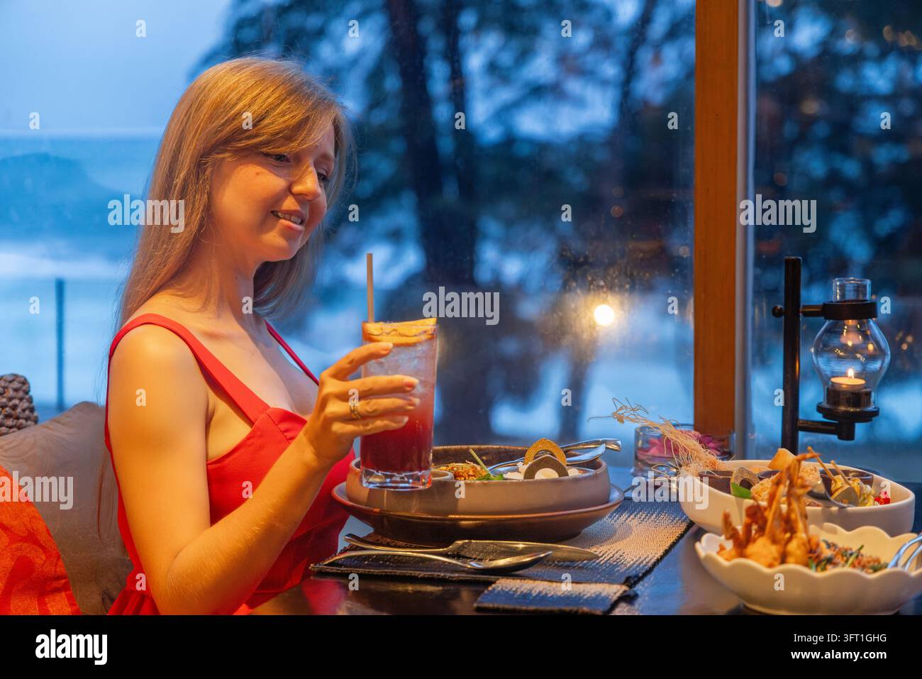 Donna in vestito rosso che assapora un cocktail rinfrescante e una cena in un ristorante di lusso in un resort tropicale. Atmosfera serale rilassante con vista sull'oceano, candele Foto Stock
