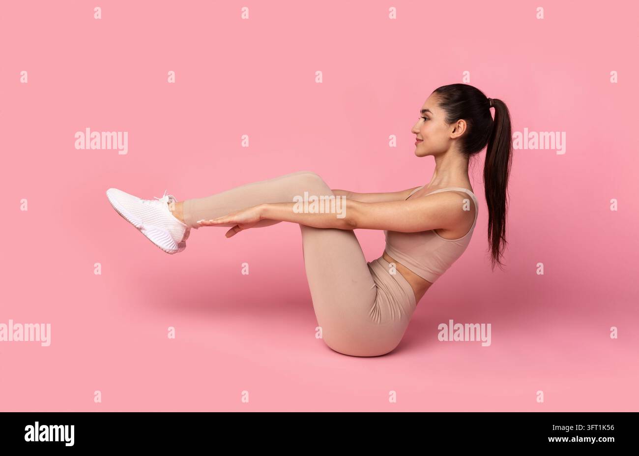 Giovane sportiva sorridente che fa addominali a terra, donna che fa crunches o sit-up con gambe sollevate e braccia dritte, lavora sugli addominali a tartaruga, studio rosa Foto Stock