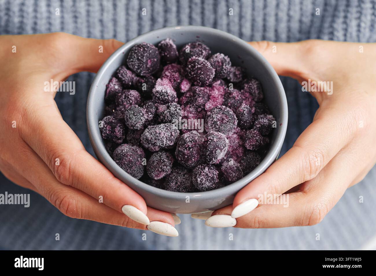 Donna che tiene una ciotola grigia di bacche scure congelate coperte di cristalli di ghiaccio. Sfondo di frutta congelata per snack sani, conservazione degli alimenti e nutrizione. Foto Stock
