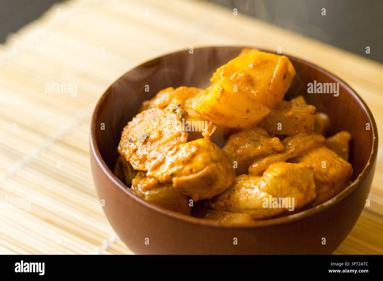 Vista laterale di curry rosso al pollo con patate in una ciotola di legno scuro su un tappeto di bambù, con vapore che sale, luce naturale soffusa Foto Stock
