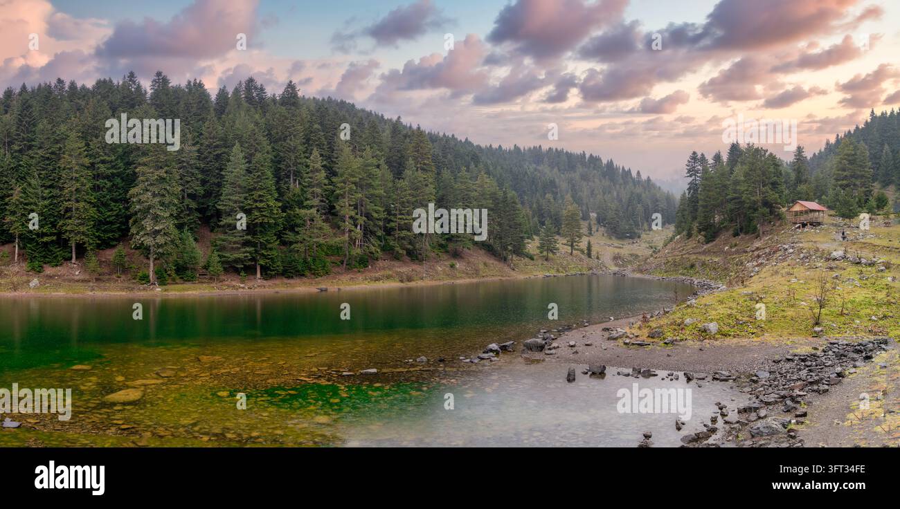 Lago di montagna ad Artvin, Turchia, circondato da una foresta di abeti, con una capanna di legno sulla riva, acqua verde chiara sopra le pietre e nuvole rosa sopra, Nero Foto Stock