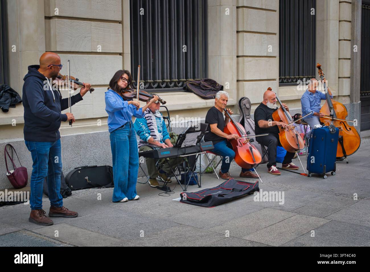 Sei musicisti di strada si esibiscono all'aperto accanto a un edificio in pietra, suonando due violini, due violoncelli, un contrabbasso e una tastiera elettronica. Foto Stock