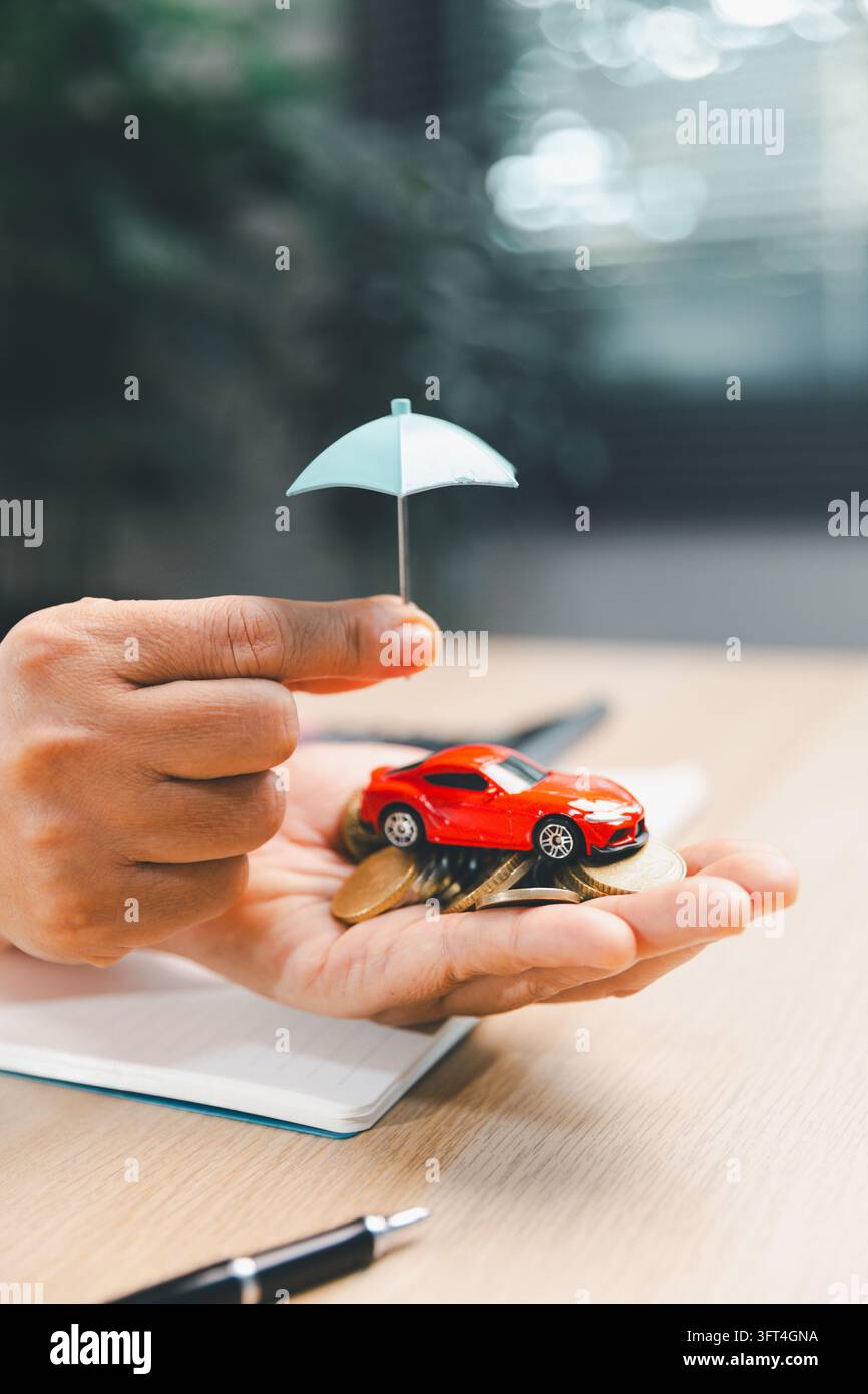 Un'auto giocattolo rossa poggia su monete nel palmo di una persona sotto un piccolo ombrello blu, con un quaderno e una penna sulla scrivania, illustrando il risparmio veicolo e f Foto Stock