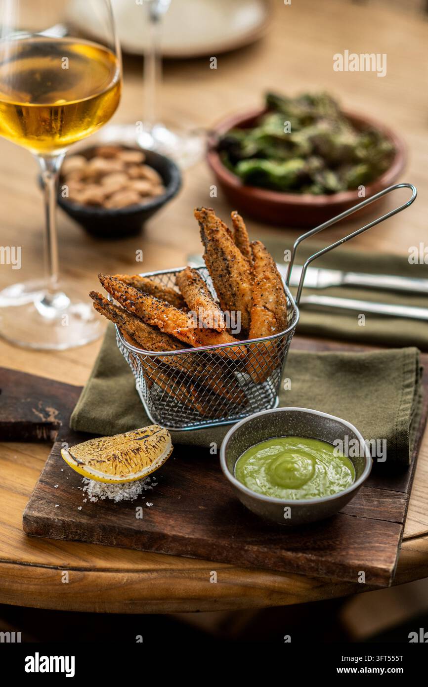 Patatine croccanti condite servite in un piccolo cestino di frittura di metallo con salsa verde da intingere e un pezzo di limone carbonizzato su una tavola di legno. Foto Stock