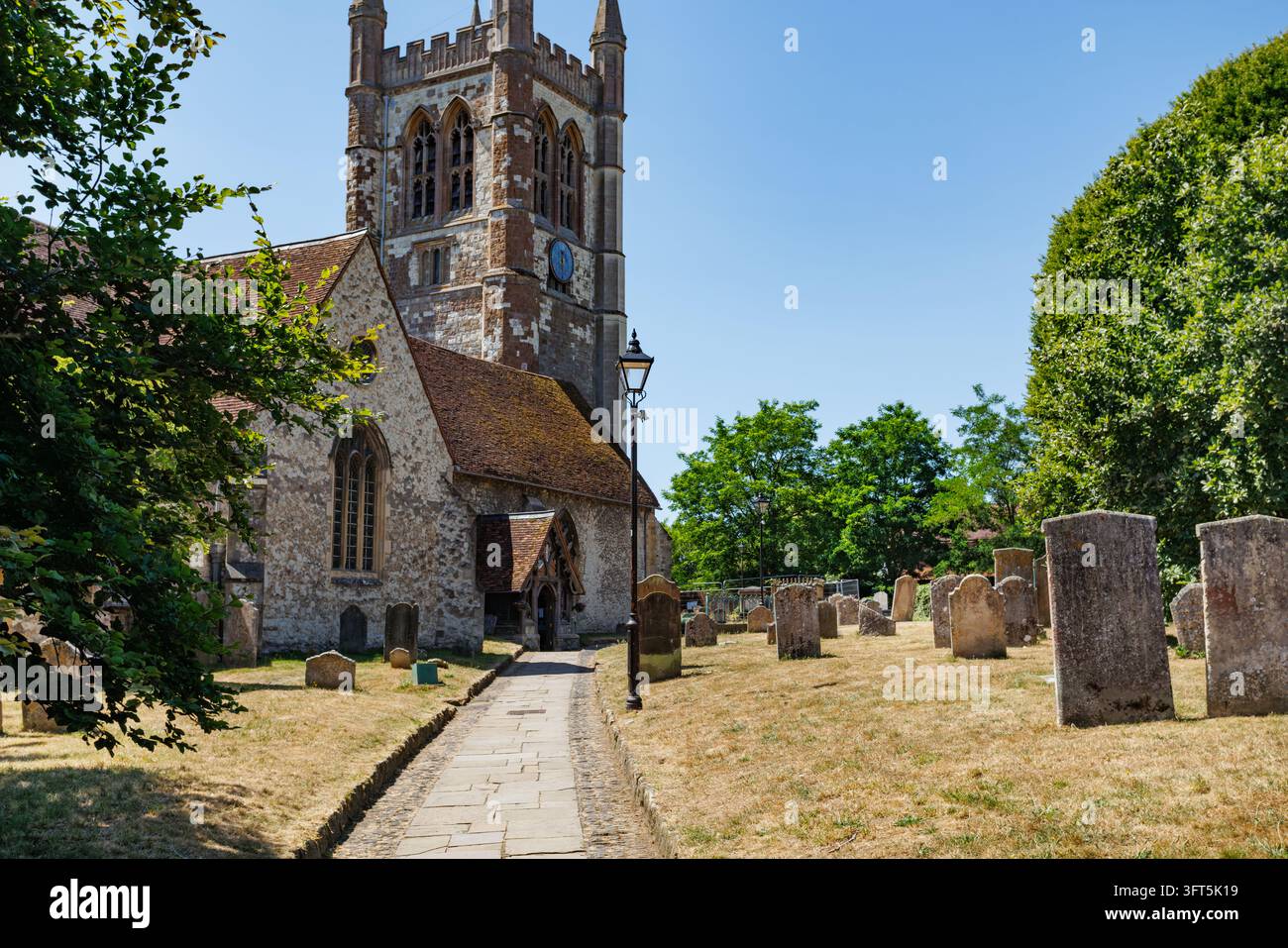 Una chiesa storica si trova accanto a un cimitero circostante con lapidi erose lungo un stretto sentiero di ciottoli, una torre alta, alberi verdi e un b Foto Stock