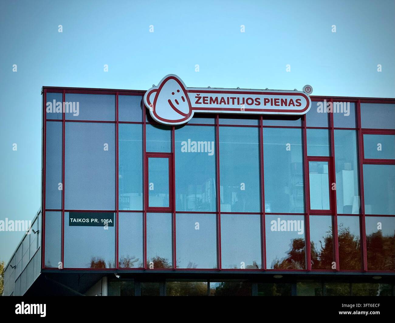 Facciata in vetro di un edificio commerciale con il logo rosso e bianco del marchio "Žemaitijos Pienas" e un cartello con l'indirizzo - Immagine stock catturata con smartphone