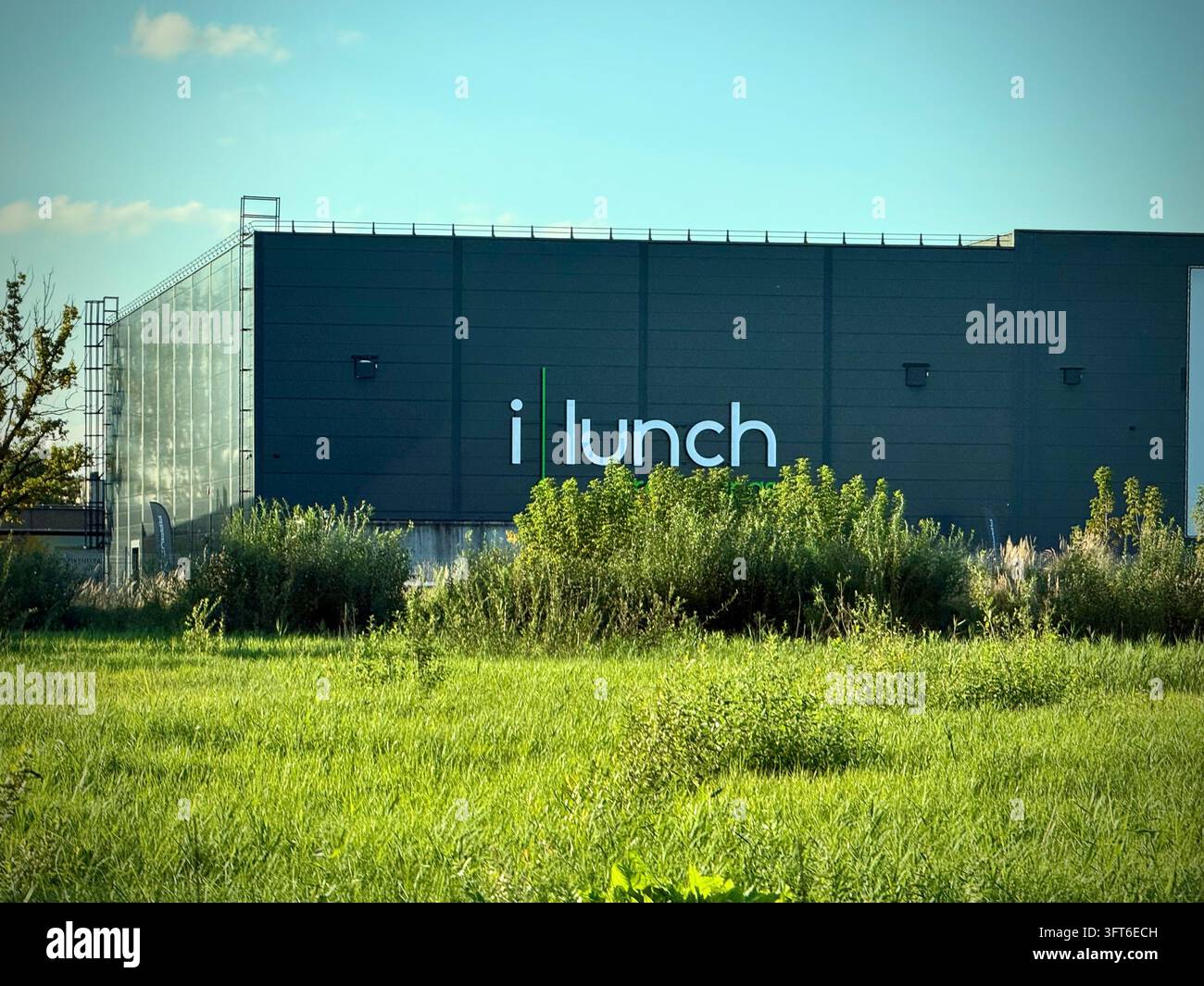 Edificio iLunch con marchio aziendale bianco, situato dietro un campo verde rigoglioso e vegetazione naturale del bordo strada sotto un cielo sereno. Foto Stock