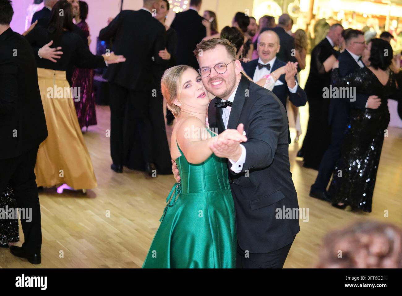 Una donna in abito da sera di raso verde balla con un uomo sorridente che indossa occhiali e un abito scuro tra altre coppie a un ballo. Foto Stock