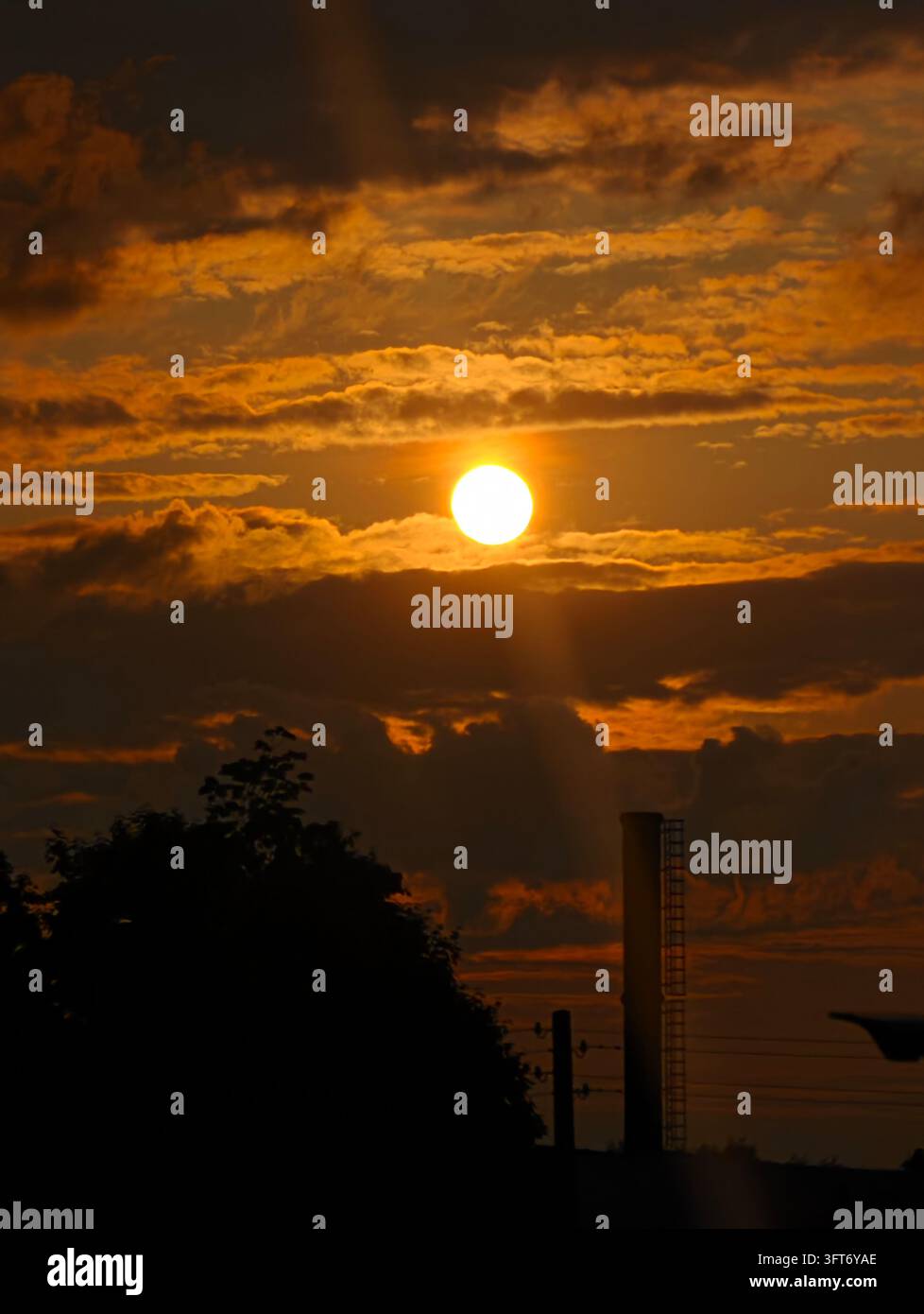 Il sole tramonta, irradiando luce dorata attraverso le nuvole. Un albero e un edificio in controluce incorniciano il cielo vibrante. Foto Stock