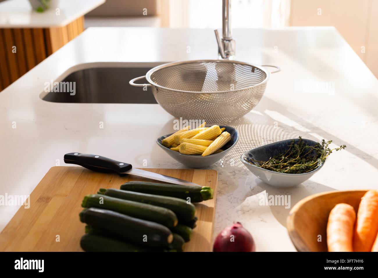 Colino in acciaio che cattura la luce del sole su un piano bianco con tavola, zucchine, coltello e ciotola di erbe Foto Stock