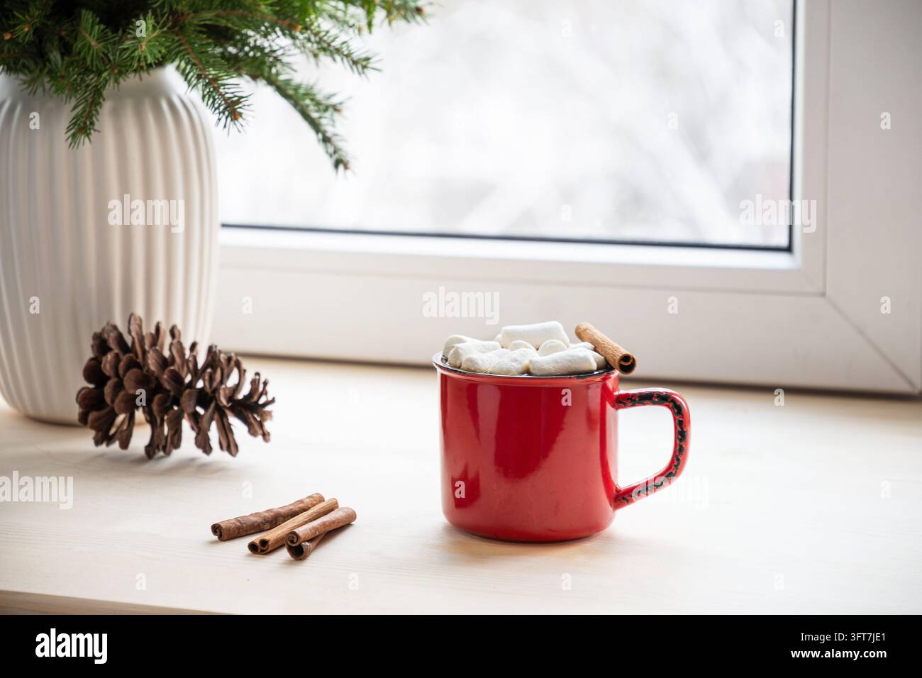 Tazza rossa con marshmallow e cannella, pigne e rami di abete sul davanzale di legno. Cioccolata calda invernale accogliente, conforto natalizio, bevanda festiva Foto Stock
