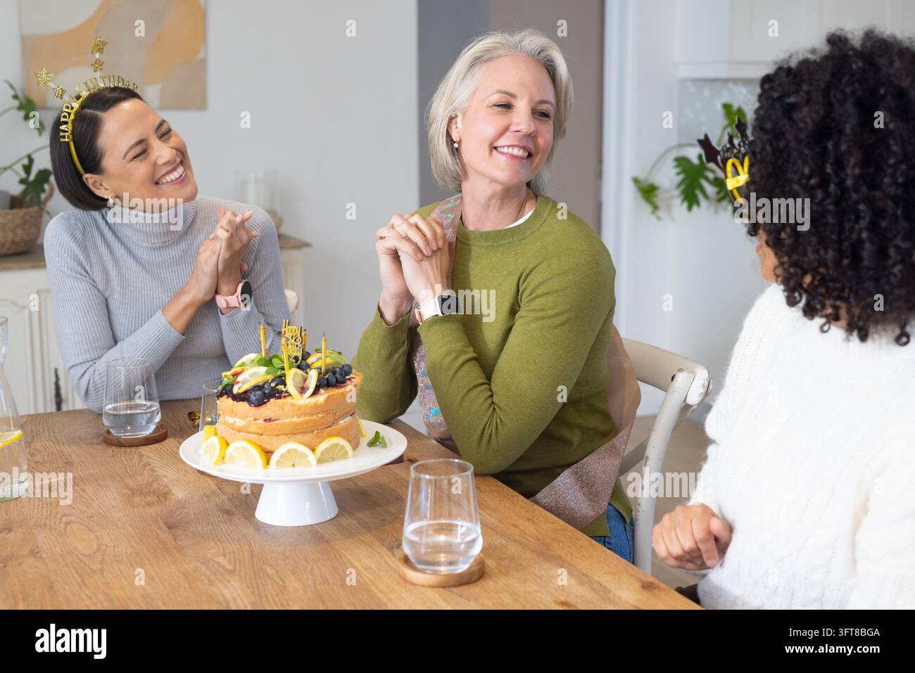 Amiche femminili sedute intorno a un tavolo di legno che celebrano il compleanno con un supporto per torte, fasce per capelli, bicchieri Foto Stock