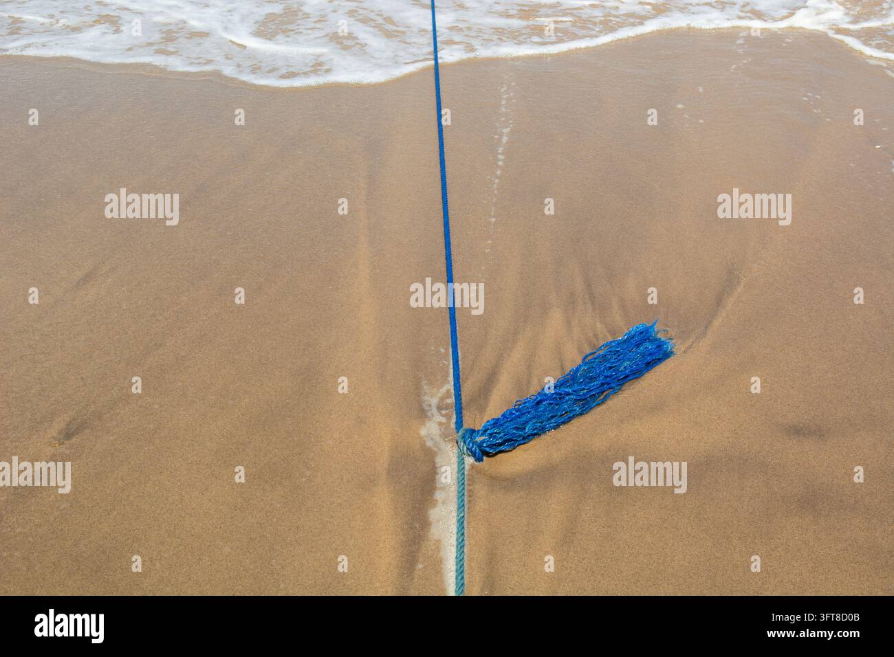 Una corda blu sfilacciata giace sulla sabbia bagnata al limite dell'acqua mentre le onde si infrangono a riva. Foto Stock