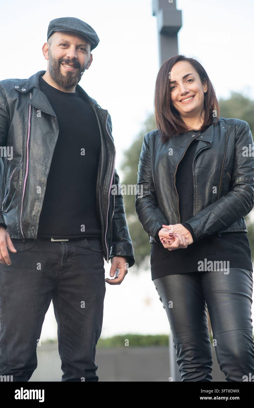Un uomo e una donna sorridenti che indossano giacche di pelle nera stanno insieme all'aperto in un contesto urbano. Foto Stock