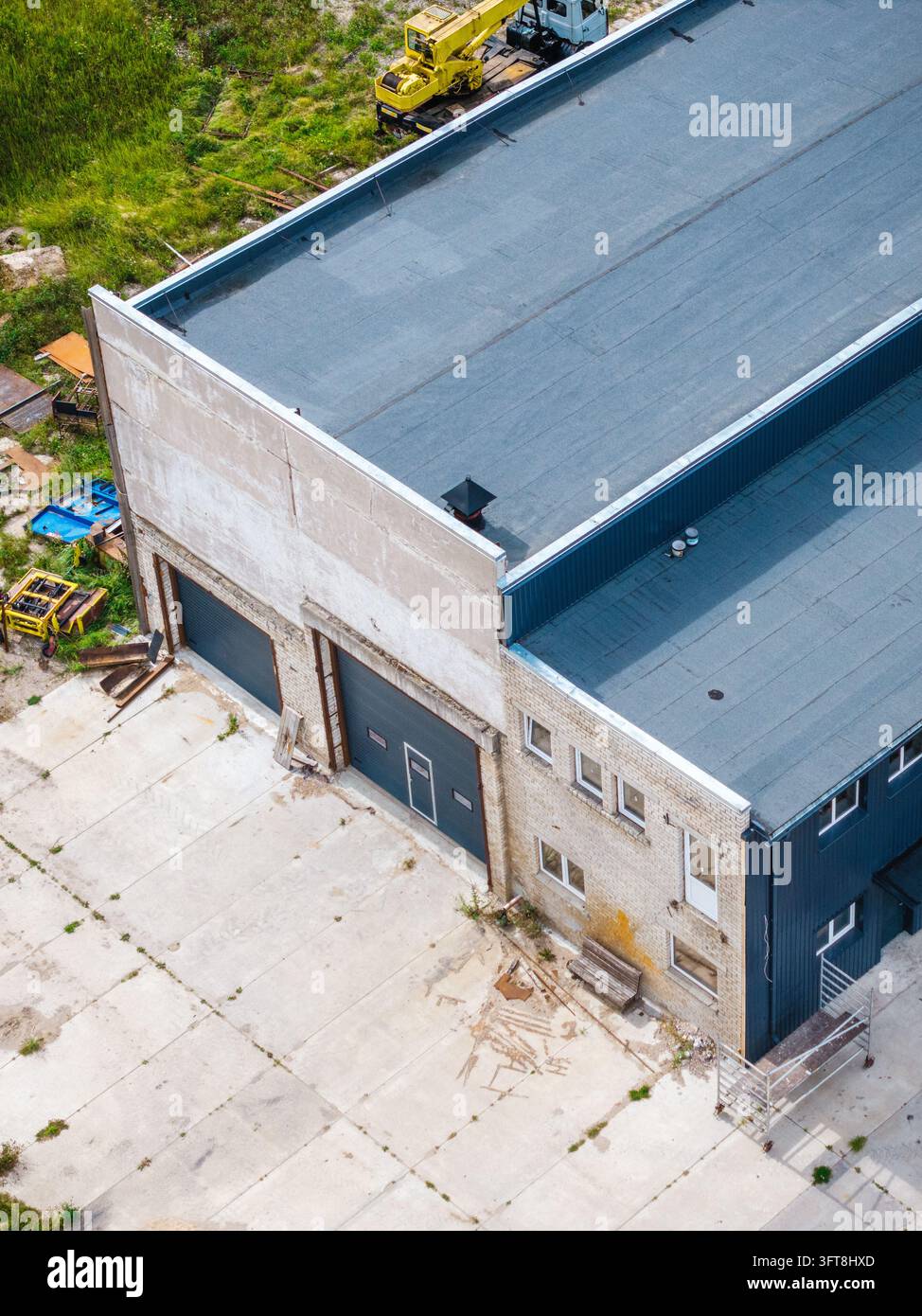 Veduta aerea di un edificio industriale con tetto piatto, grandi porte del garage e una gru gialla parcheggiata nel cortile di cemento a Zlekas, Ventspils Munici Foto Stock