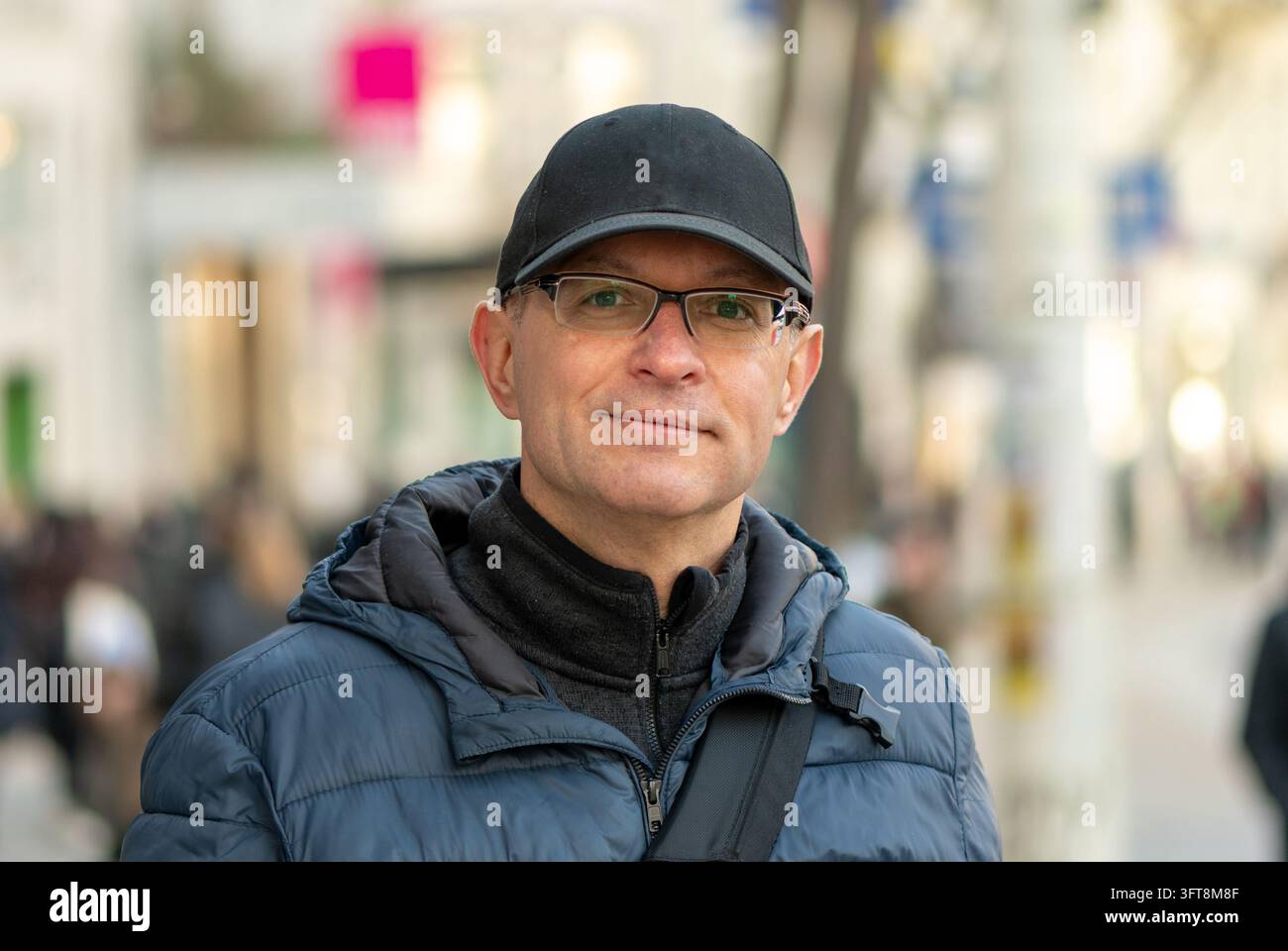Un uomo di mezza età con un cappello nero e occhiali si trova su una strada cittadina sfocata. Foto Stock