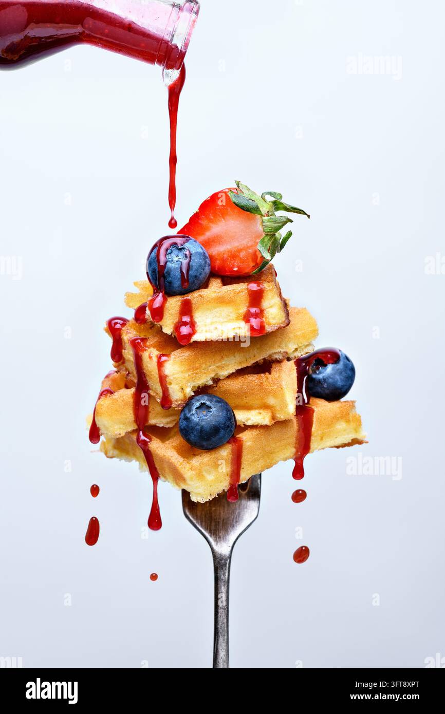 Pila di waffle dorati guarniti con fragole fresche e mirtilli, colati con sciroppo di bacche rosse che cola da una bottiglia di vetro Foto Stock