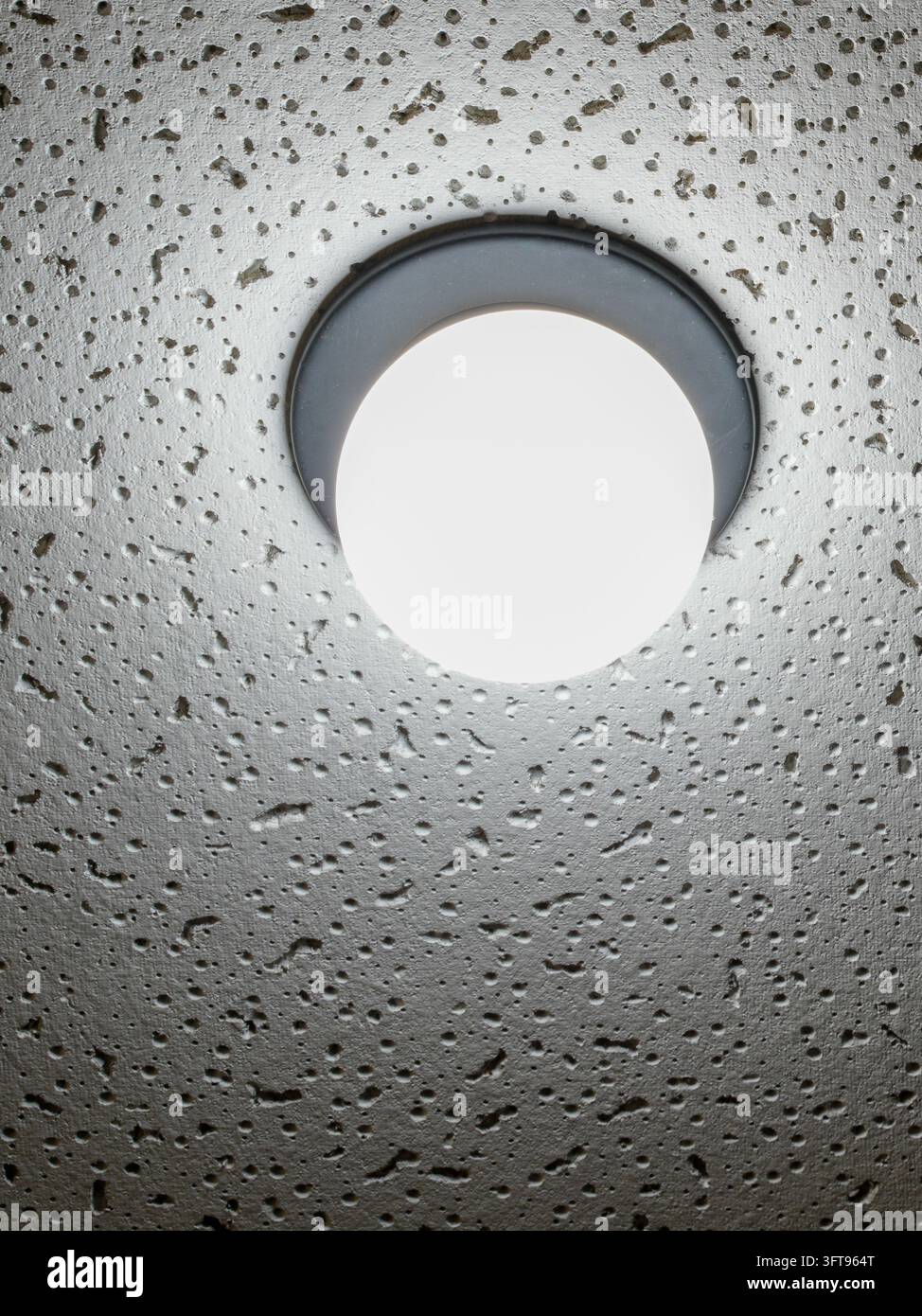 Primo piano di una lampada da soffitto a LED rotonda luminosa montata su un soffitto in cemento ruvido e poroso. L'illuminazione moderna contrasta con una superficie industriale ruvida. Foto Stock