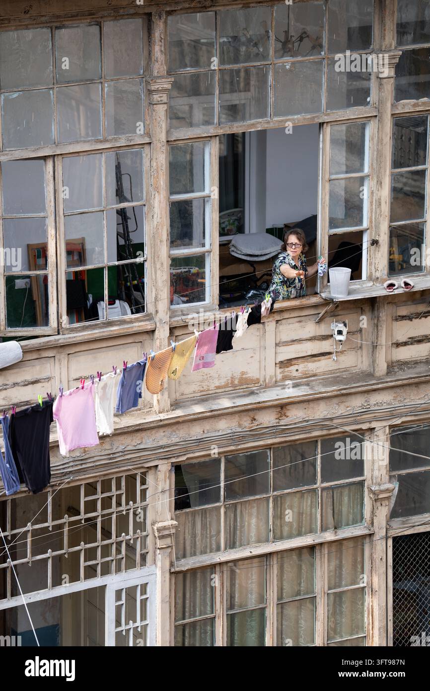 Anziana che stende il bucato dalla finestra in una vecchia casa di legno storica a Tbilisi, Georgia. Foto Stock