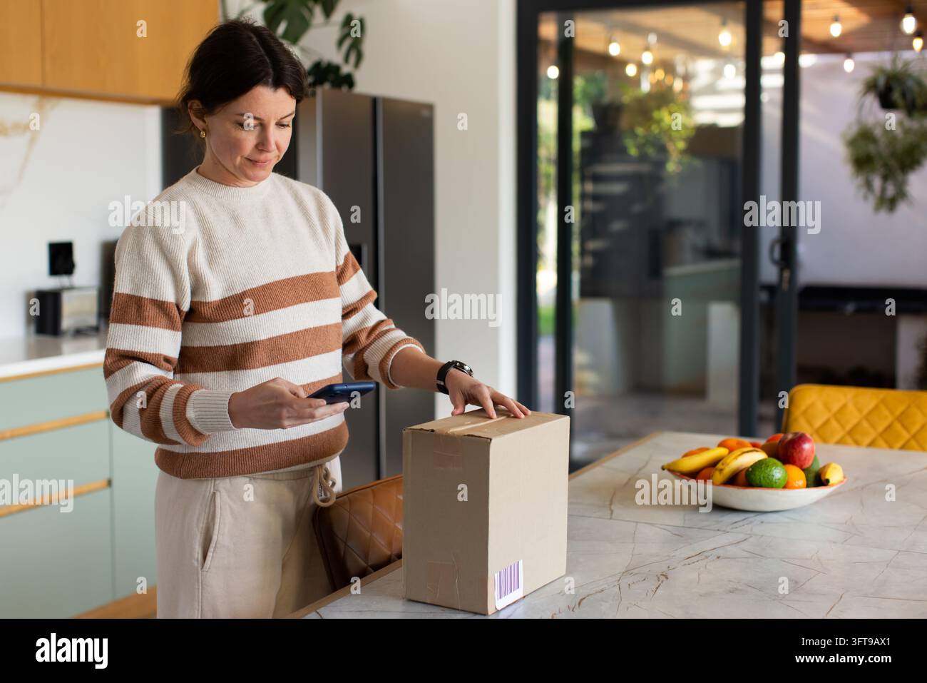 Donna che tiene uno smartphone mentre sostiene una scatola di cartone sull'isola della cucina, scansionando un codice a barre Foto Stock
