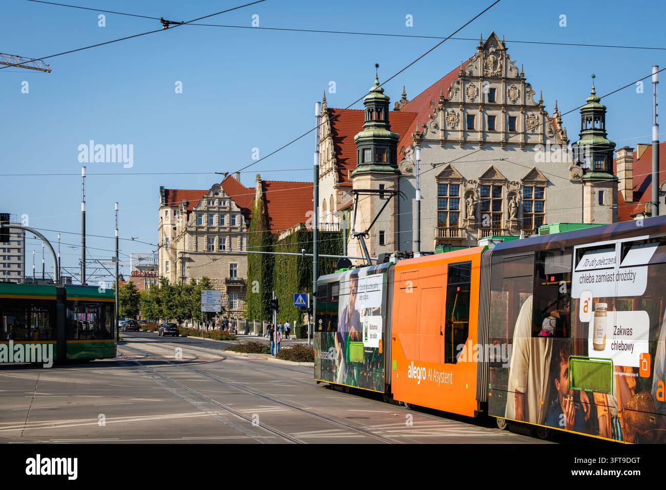 Una veduta cittadina diurna soleggiata a Poznan, in Polonia, che mostra tram moderni di trasporto pubblico arancioni e verdi sui binari davanti all'architettura neo-renaissance storica Foto Stock