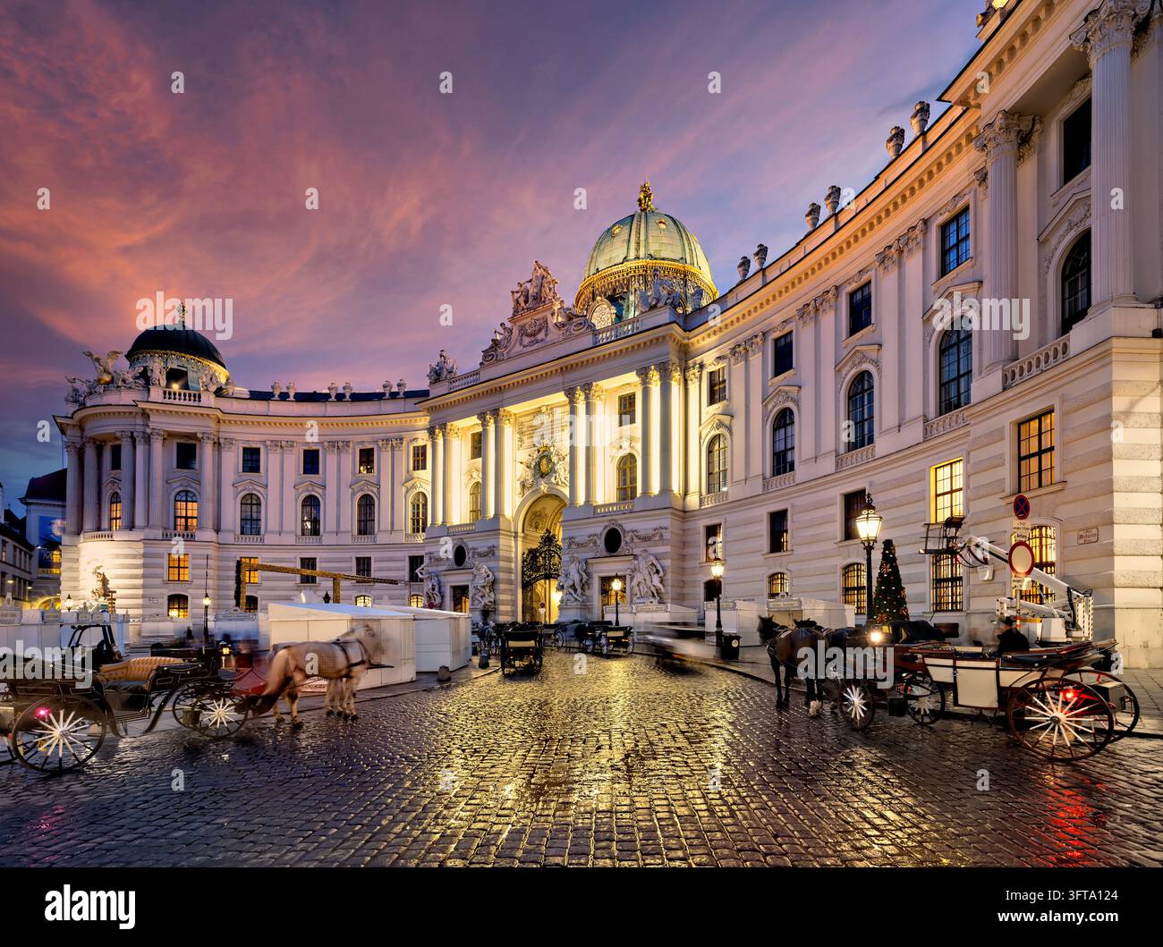 Carrozze trainate da cavalli attendono sui ciottoli bagnati di Michaelerplatz davanti all'ala illuminata di San Michele del Palazzo Imperiale al crepuscolo invernale Foto Stock
