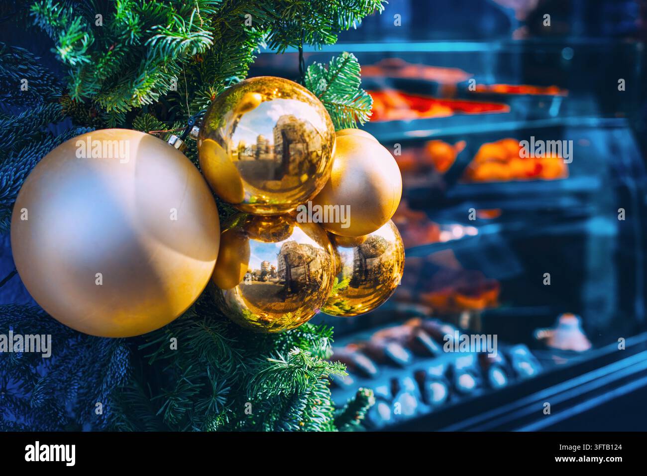 Fuori da una vetrina del negozio con una esposizione di pasticceria, delle palline di Natale sono appese a rami di abete. Gli edifici si riflettono sulle superfici lucide degli ornamenti Foto Stock