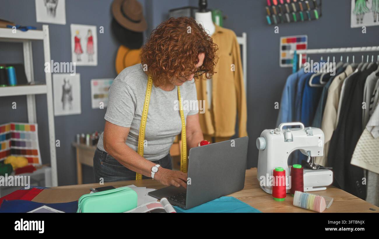 Donna che digita su un laptop con le mani sulla tastiera, metro intorno al collo e macchina da cucire nelle vicinanze in studio; concentrazione artigianale. Foto Stock