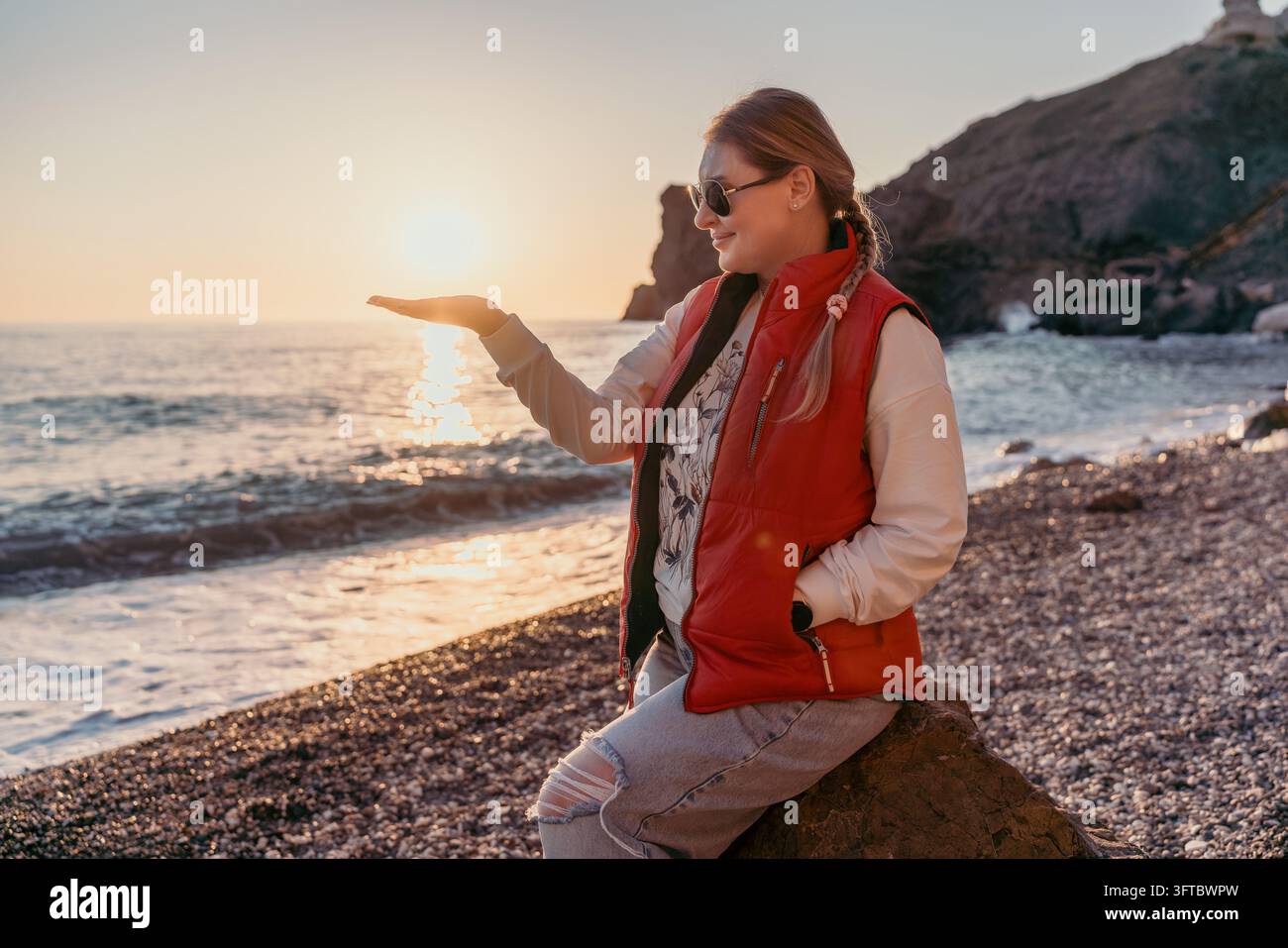 Donna sorridente sulla spiaggia al tramonto, indossa un giubbotto rosso, tiene il sole che tramonta sul palmo mentre è seduta su una riva rocciosa Foto Stock