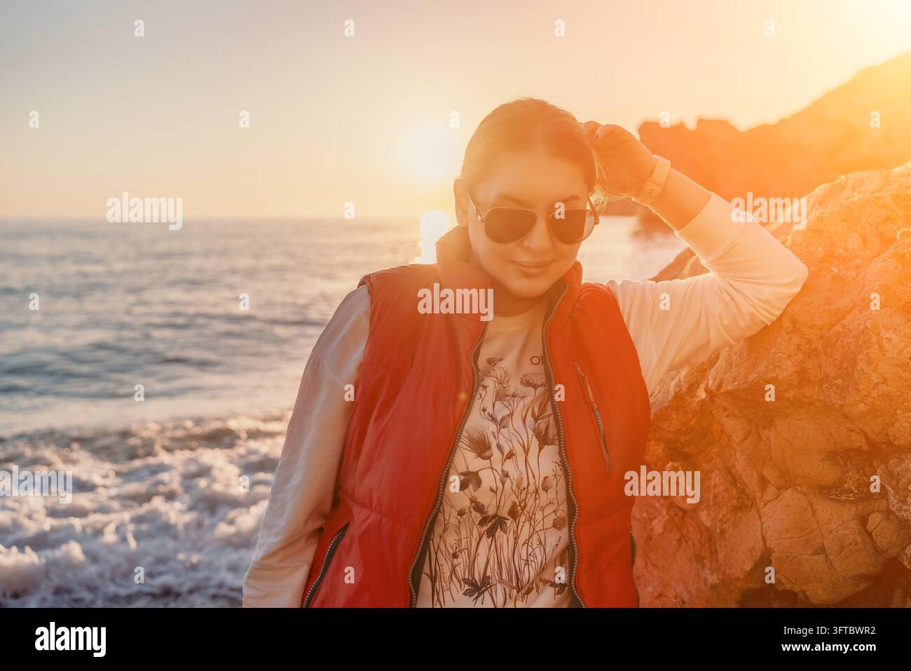 Donna tramonto spiaggia donna con un giubbotto rosso appoggiata su scogli vicino all'oceano all'ora dorata con flare della lente Foto Stock