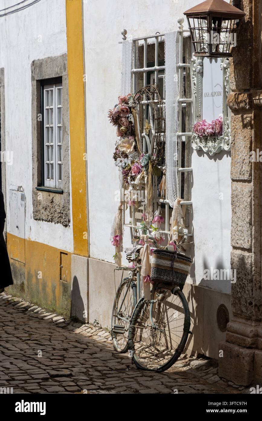 Obidos, Portogallo - 9 maggio 2026: Bici vintage e ghirlanda floreale decorano la facciata bianca tradizionale di una casa nel villaggio medievale. Foto Stock
