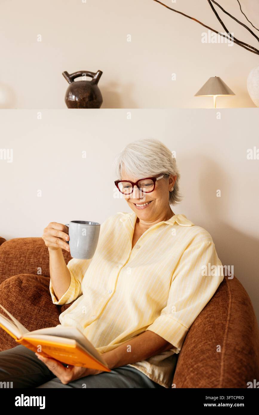 Donna anziana che si gode il tempo libero sul divano, legge un libro e tiene una tazza di caffè Foto Stock