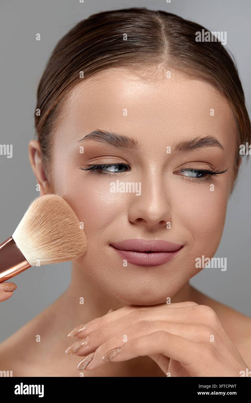 Bella donna applica un pennello trucco soffice sulla guancia mostrando una pelle liscia e radiosa e un sorriso sottile. Giovane donna usa il pennello per un elegante Foto Stock