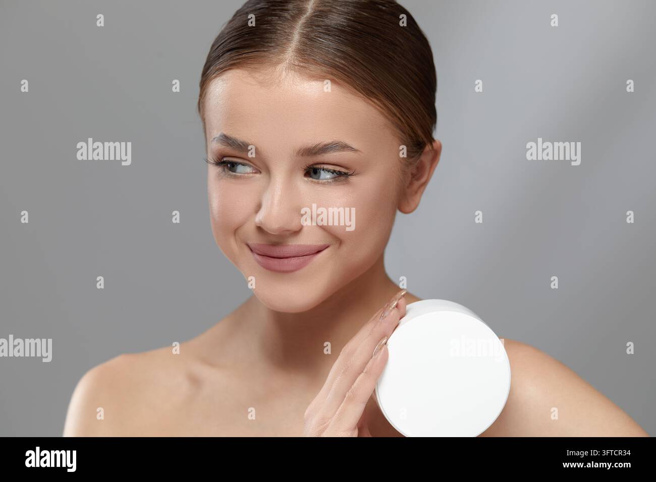 Donna attraente tiene un barattolo di crema bianco accanto al viso mostrando una pelle liscia e luminosa e un sorriso sottile. Donna di bellezza presenta i prodotti per la cura della pelle Foto Stock