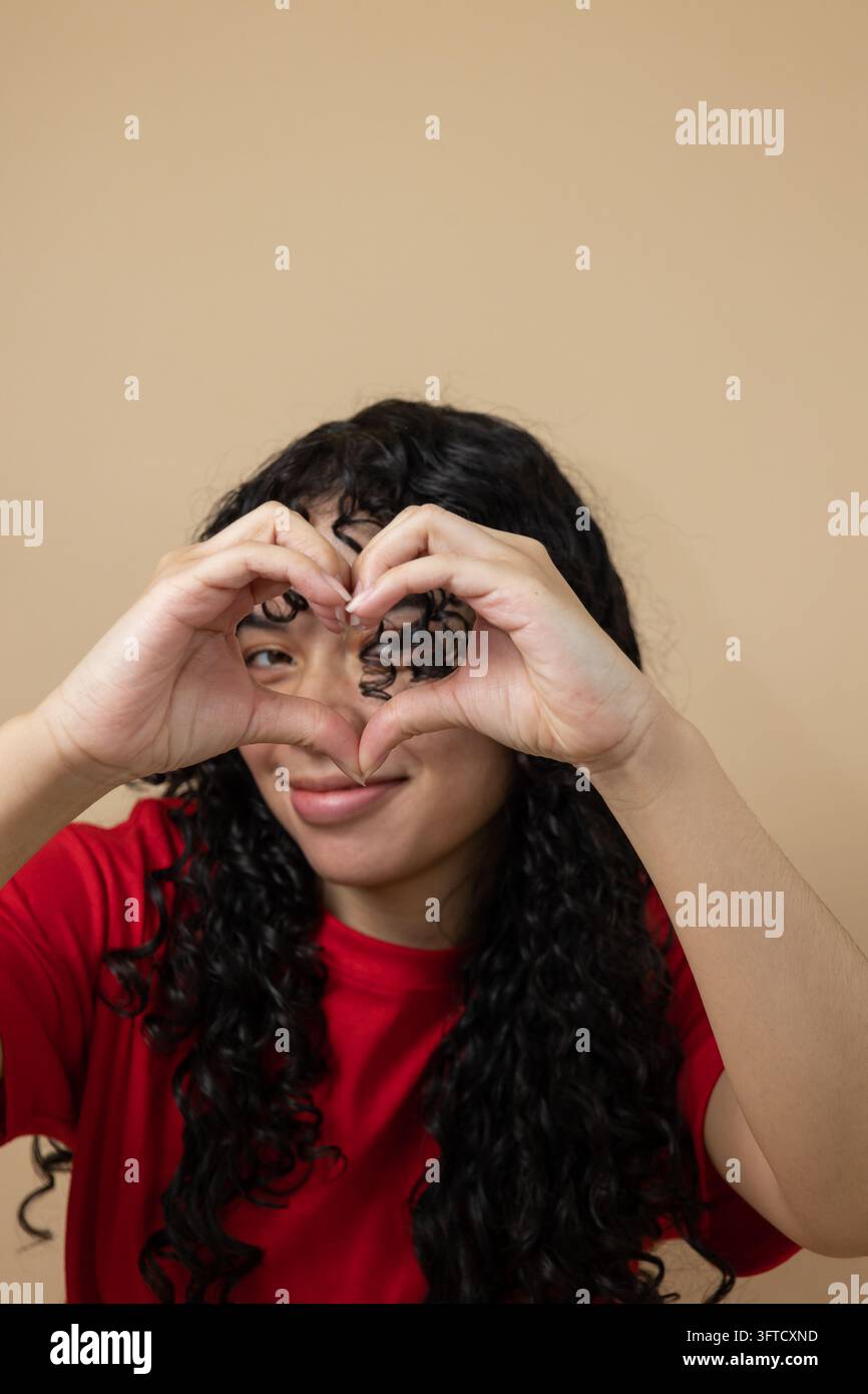 Donna latina allegra in una camicia rossa che forma un cuore con le mani e guarda attraverso di esso su uno sfondo marrone chiaro con spazio per il testo concetto di amore Foto Stock