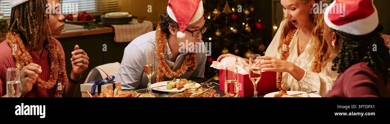Intestazione di amici diversi che aprono un regalo di Natale durante la cena delle feste Foto Stock