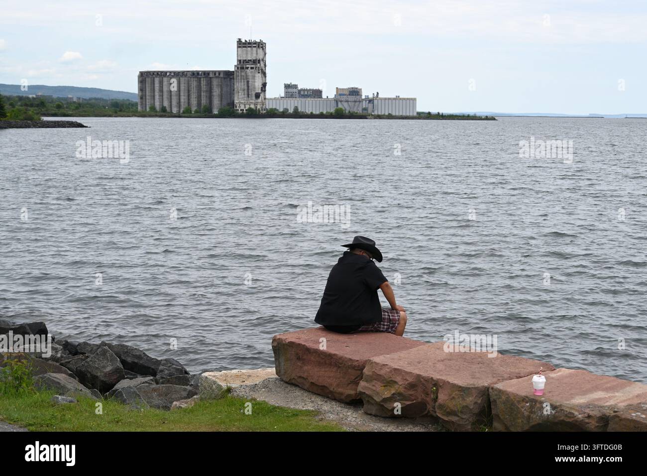 Thunder Bay, Ontario, Canada – 01 luglio 2026: Un uomo è seduto sul lungomare di Thunder Bay con l’Antico Elevatore per Cereali sullo sfondo. Foto Stock