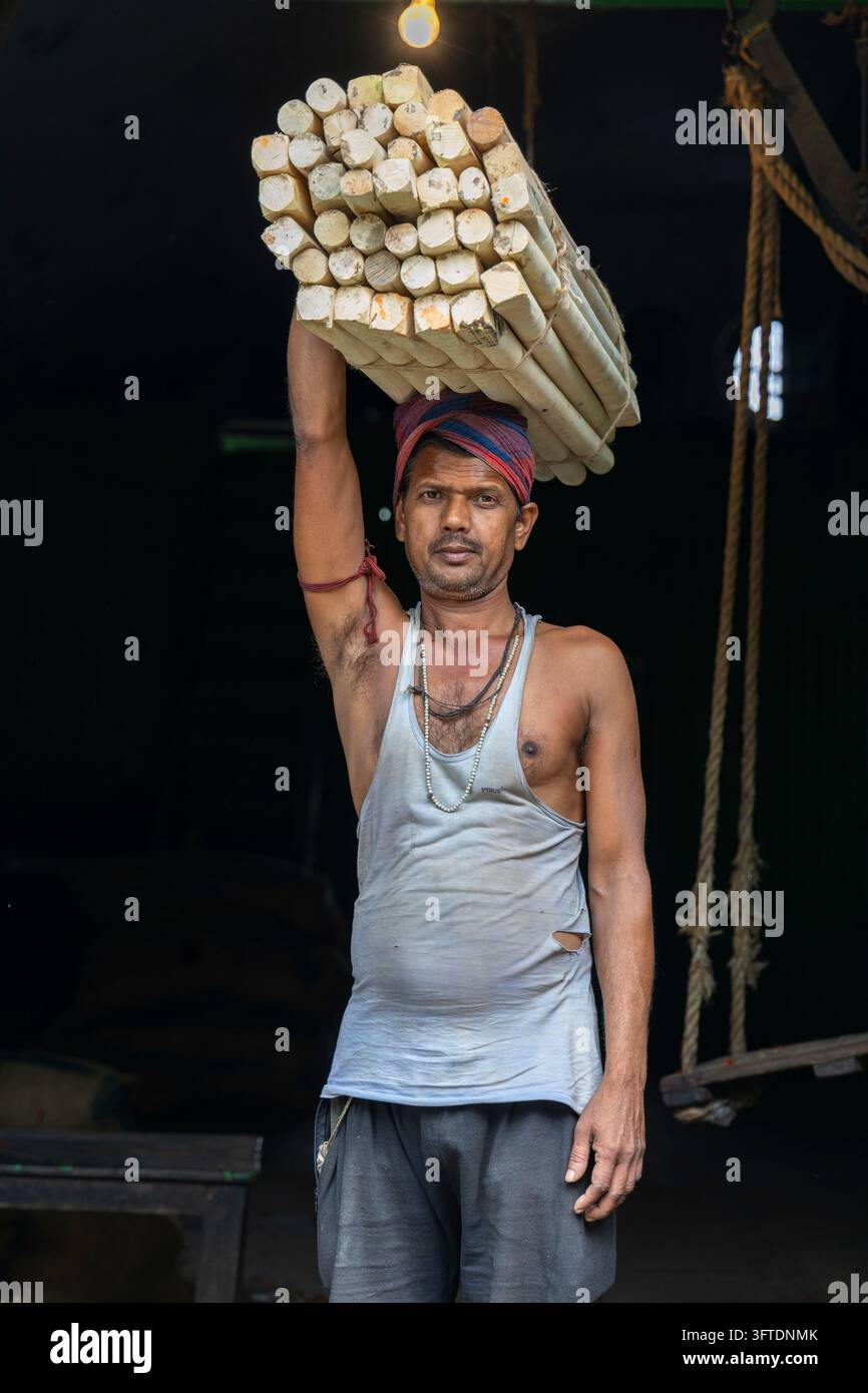Un portiere con un gilet grigio e un panno rosso bilancia un grosso mazzo di bastoncini di legno tagliati sopra la testa, Bengala Occidentale, Howrah, India Foto Stock