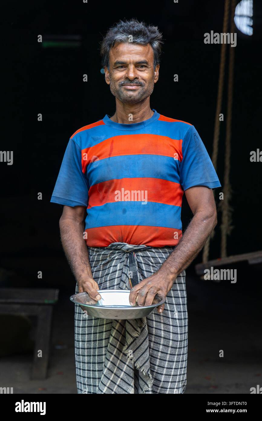 Un lavoratore sorridente con capelli grigi scompigliati si trova con entrambe le mani su un piatto di metallo davanti a un interno scuro, Bengala Occidentale, Howrah, India Foto Stock